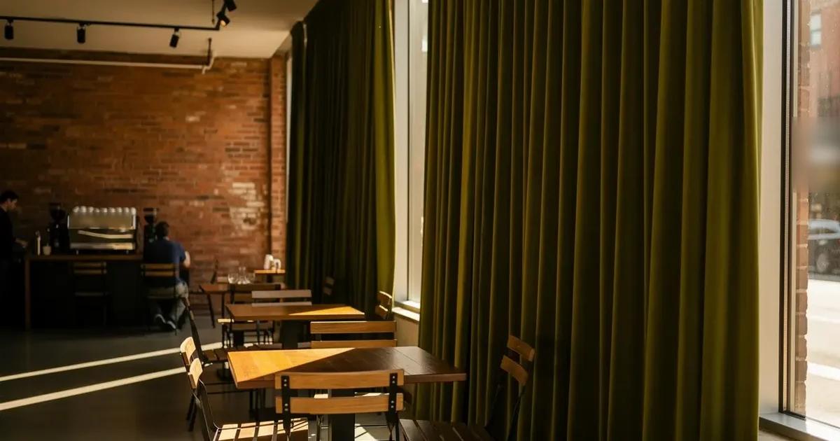 Olive velvet curtain gathered along the tall street-facing windows of a brick-walled café, wooden tables, metal chairs and an espresso machine at the back of the room