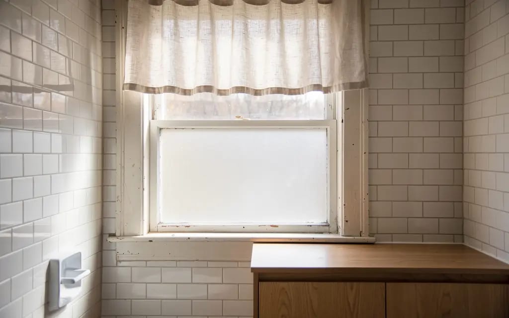 Bathroom window with a short linen cafe curtain mounted above the frame