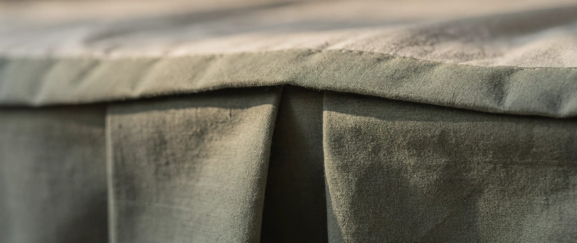 Crisp box pleat of a sage-green cotton bed skirt, with deep shadow inside the fold.