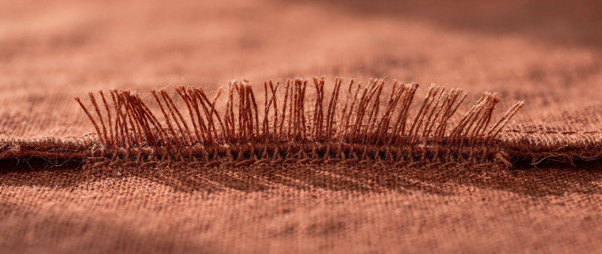 Frayed edge of a terracotta linen swatch, loose warp threads fanning out into a fringe.