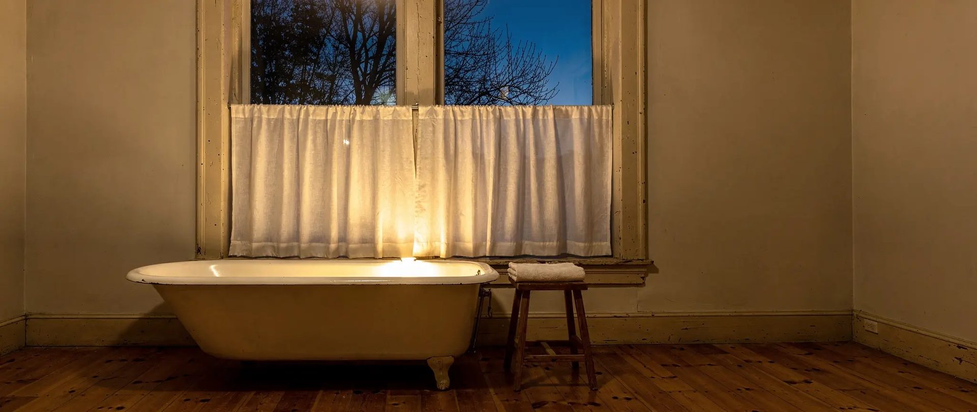 Rideau bistro en lin blanc sur la moitié basse d'une fenêtre de salle de bain, le soir, au-dessus d'une baignoire sur pattes, le haut de la vitre laissé dégagé