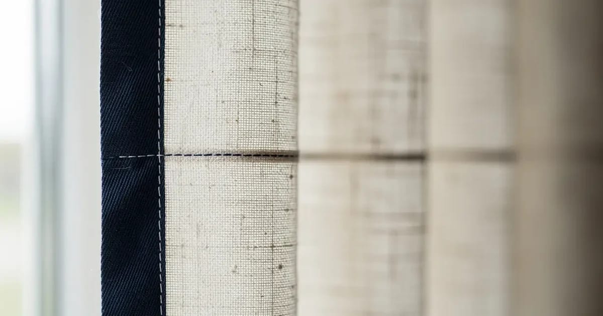 Détail du bord menant d'un rideau en lin écru orné d'une bande contrastante en coton bleu marine large de huit centimètres, couture nette visible de près, plis réguliers, lumière naturelle latérale dans un atelier de Sherbrooke