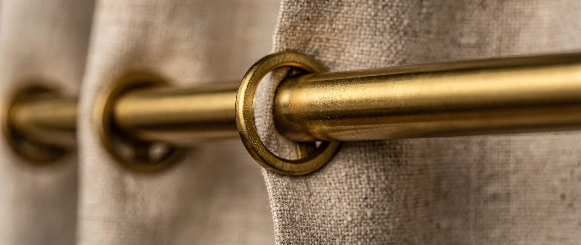 Brushed brass rod with rings gripping stone-beige linen, shown in close-up.