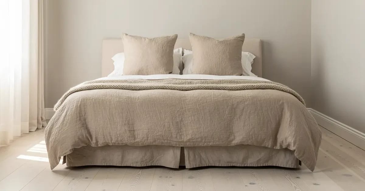 Lit double dans une chambre de Sherbrooke habillé d'une housse de couette en lin lavé beige tombant également des deux côtés jusqu'au cache-sommier, deux taies européennes appuyées contre la tête de lit, lumière naturelle du matin par une fenêtre habillée de voilage
