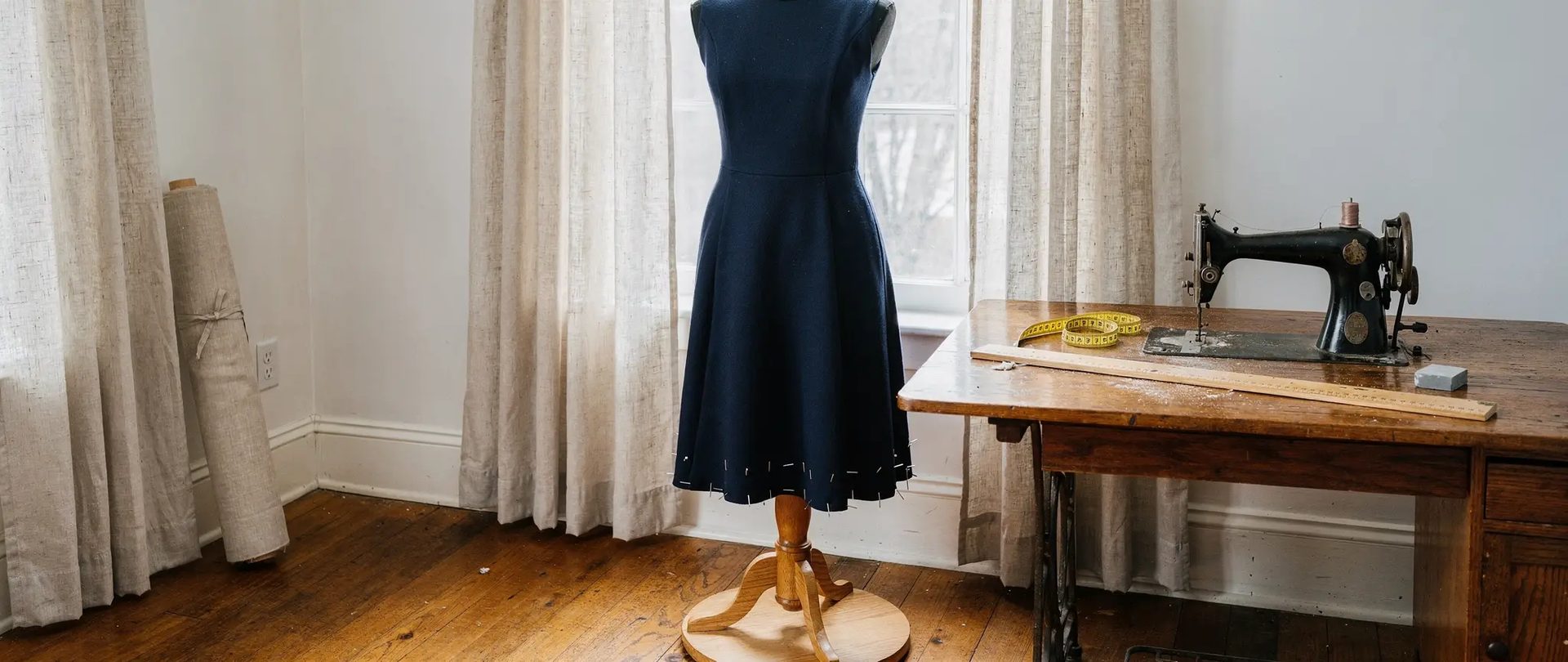 Clothing alterations in a sewing atelier: a navy wool dress on a form, its hem pinned, and linen curtains at the window.