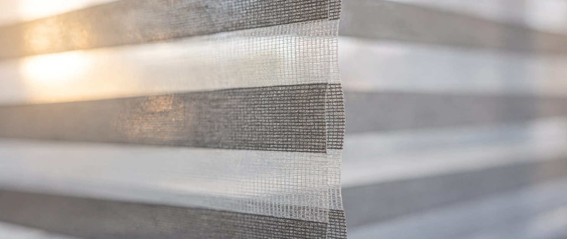 Alternating sheer and opaque bands of a grey and white zebra blind, seen from the side.