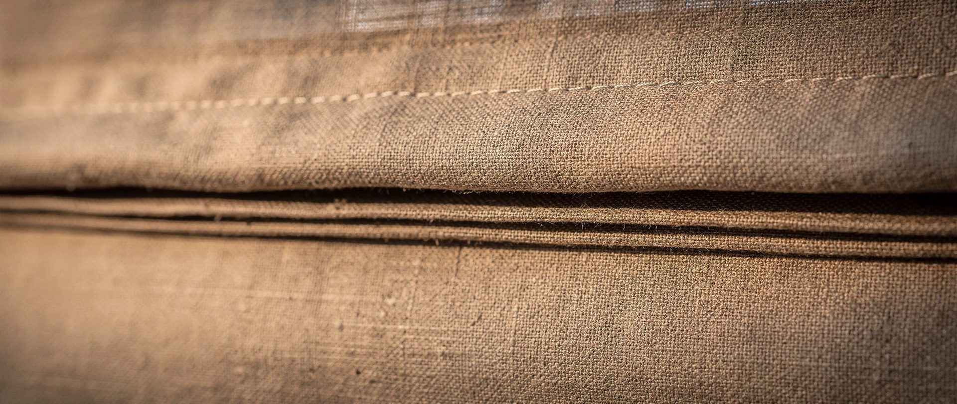Flat folds of a mushroom-coloured linen Roman shade with a crisp fold and visible stitching.