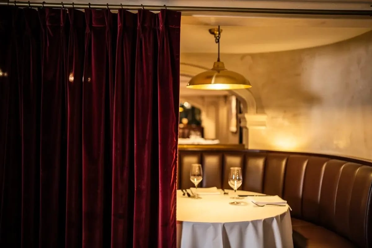Plis d'un rideau de velours bordeaux au premier plan, à demi tiré devant une alcôve; une table dressée pour deux et une banquette de cuir sous une suspension en laiton.